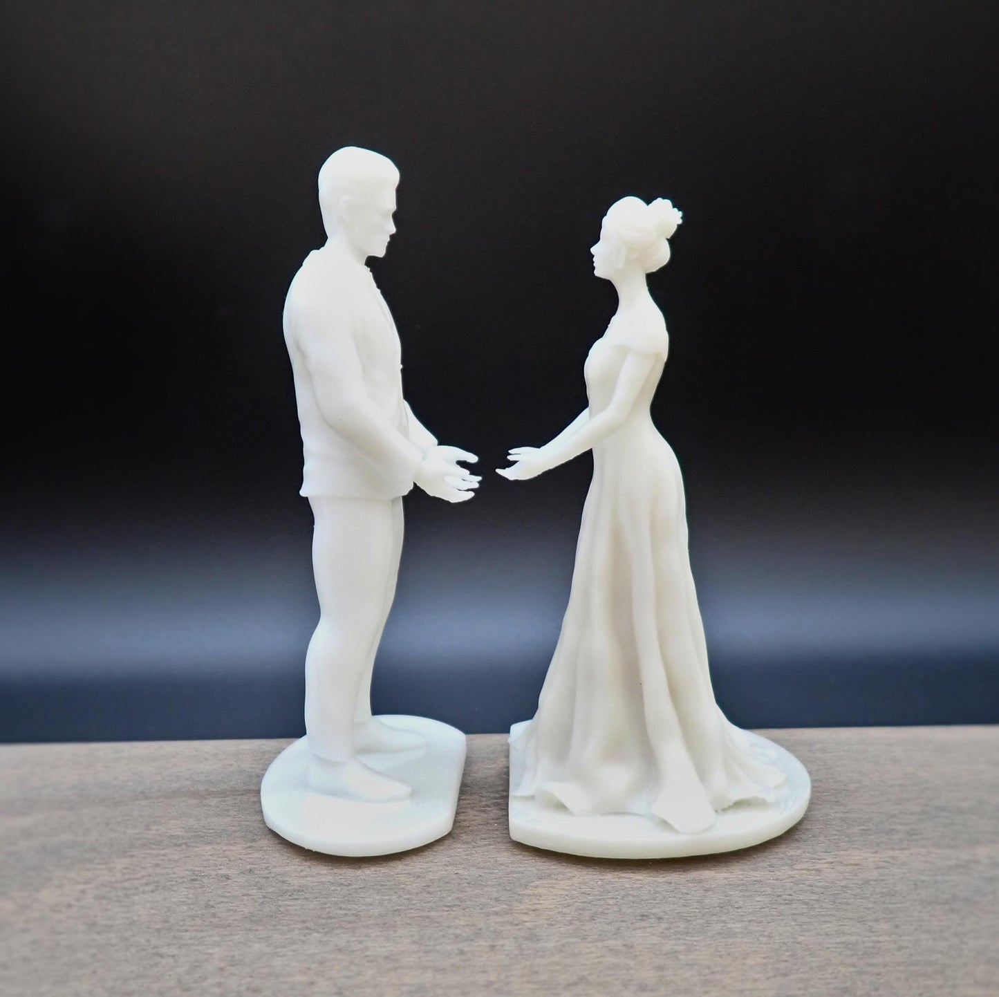 A pair of unpainted cake toppers depicting a bride and groom in a romantic pose, standing on a wooden surface.